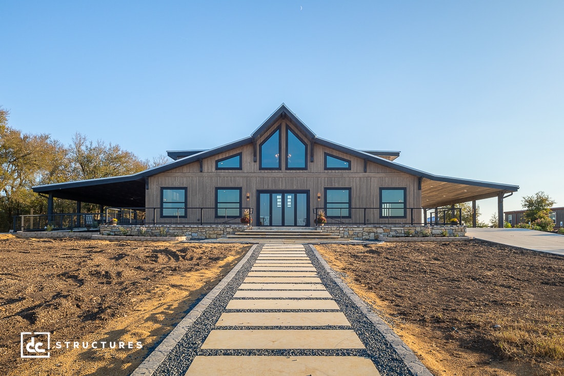 A modern barn-style building with large windows and a covered porch, front view. Stone walkway leads through dry, sparse landscape.