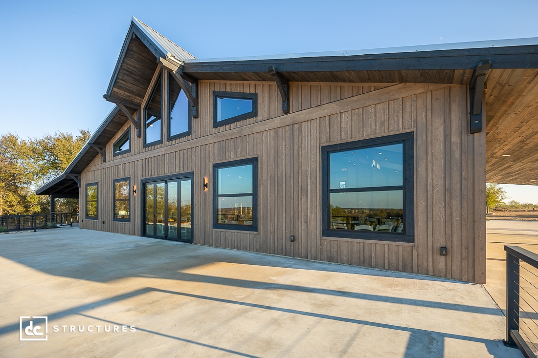 A modern barn-style building with large windows and wooden siding, high angled roof, and a spacious concrete patio under a clear sky.