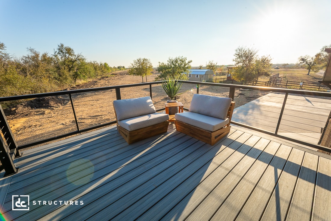 A spacious outdoor deck with two cushioned wooden chairs and a potted plant overlooks a sunny rural landscape. Black metal railings enclose the deck.