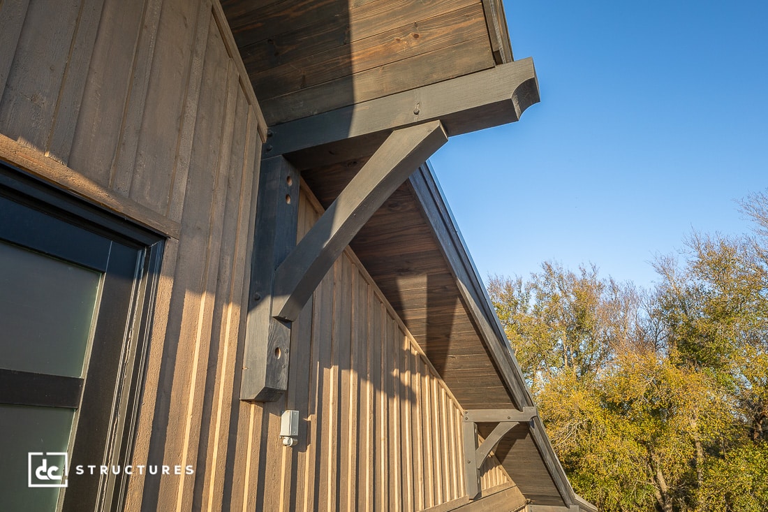 Close-up view of a wooden building’s exterior, focusing on angled support beams under a dark-stained roof eave against a blue sky, with trees and sunlight in the background.