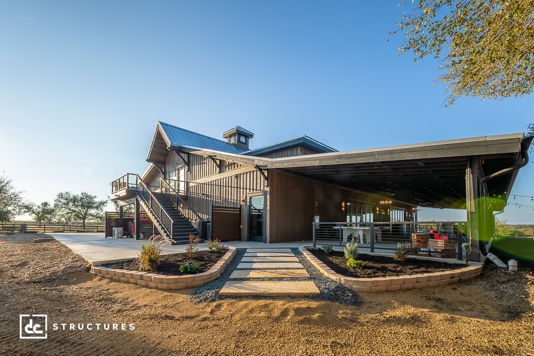 A modern barn-style building with a metal roof, outdoor staircase, covered patio with seating, landscaped garden beds, clear blue sky.