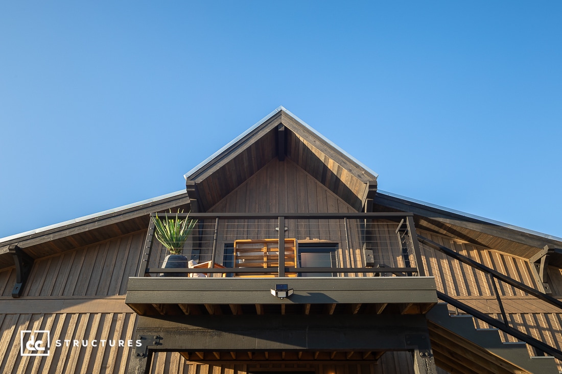 A modern wooden building with a balcony, featuring a table, chairs, and a potted plant under a clear blue sky. The structure has dark trim and vertical wood paneling.