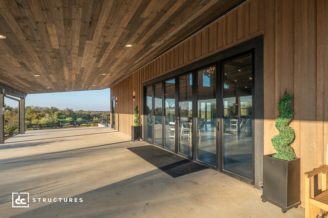 A spacious covered patio with a wood-paneled ceiling, glass doors, and potted plants outside a modern building overlooking a scenic landscape.