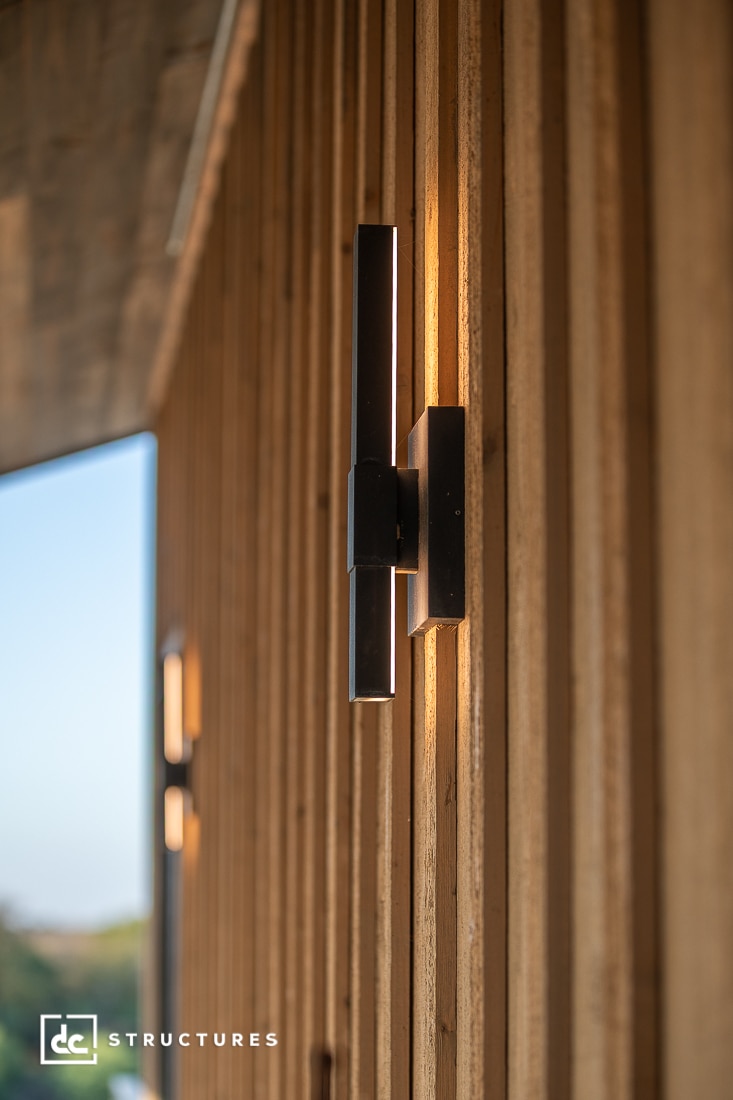 A modern black wall sconce is mounted on vertical wooden siding, emitting warm light. The background is softly blurred.
