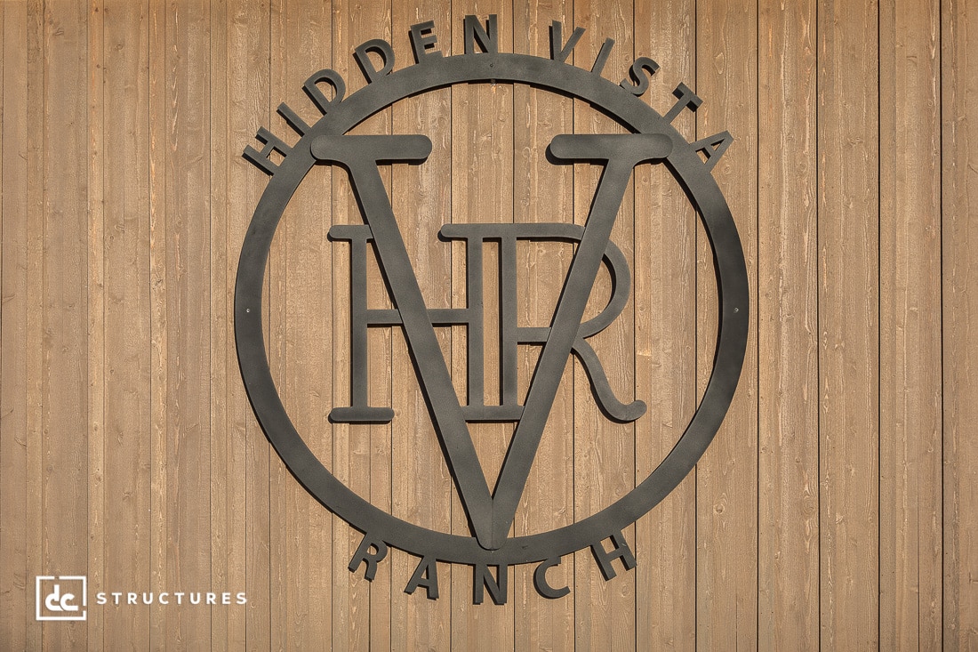 A metal sign with the letters HVR inside a circle and the words Hidden Vista Ranch above and below, mounted on a wooden wall.