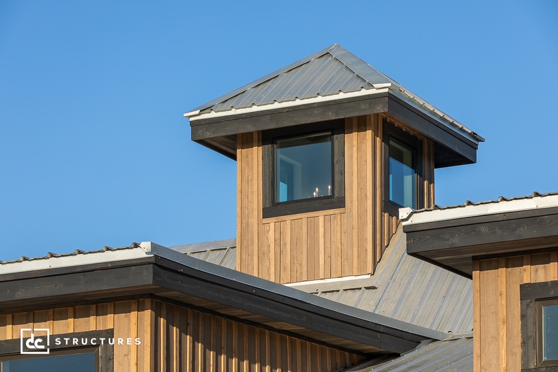 A close-up of a modern wooden building with a metal roof, featuring a small tower with large windows under a clear blue sky.