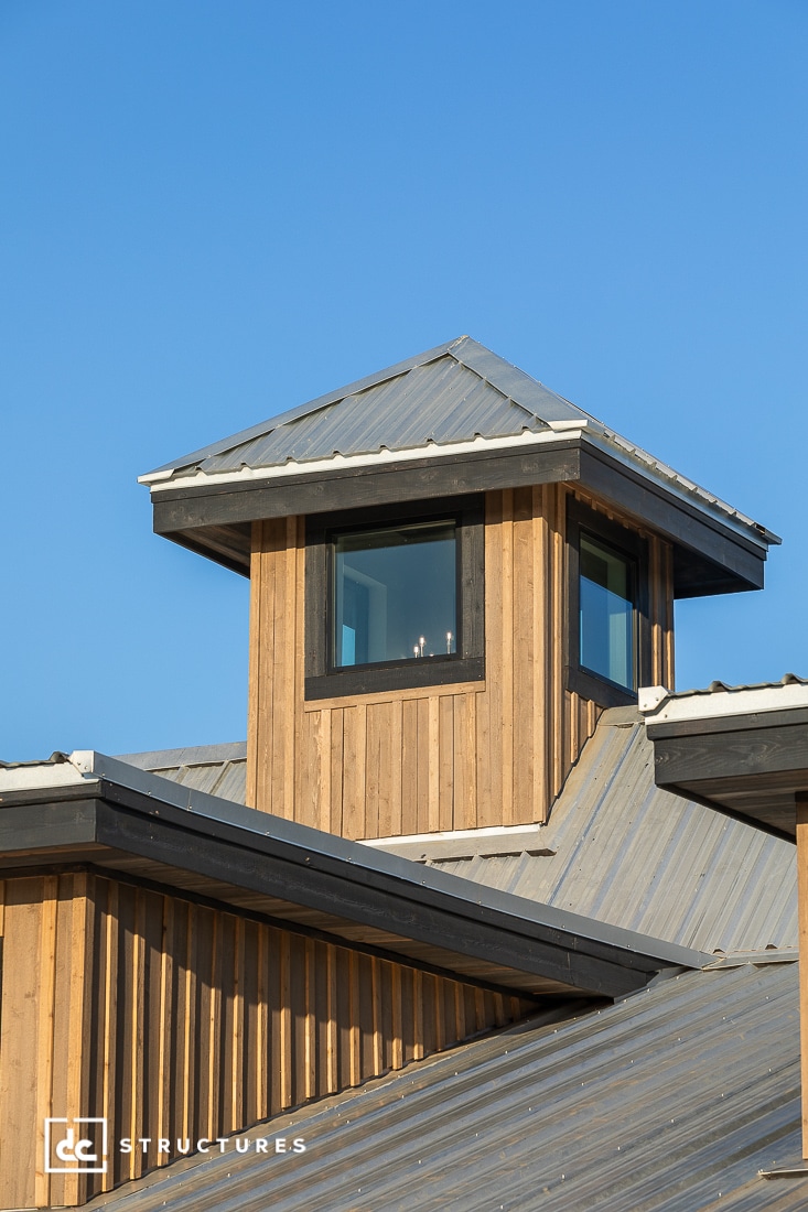 A wooden cupola with large windows and a metal roof sits atop a modern building with matching metal and wood siding under a clear blue sky.
