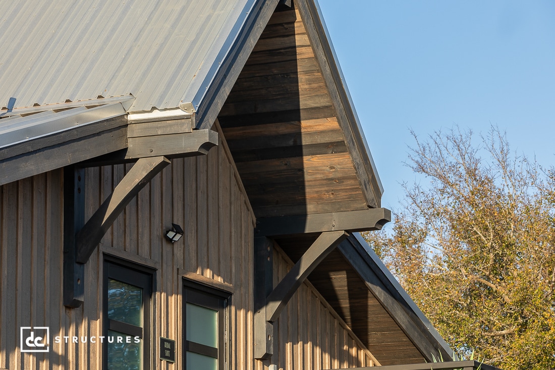 Close-up of a modern barn-style building with a sloped metal roof, wood siding, and exposed beams. Sunlight highlights textures. Trees and blue sky are visible in the background.