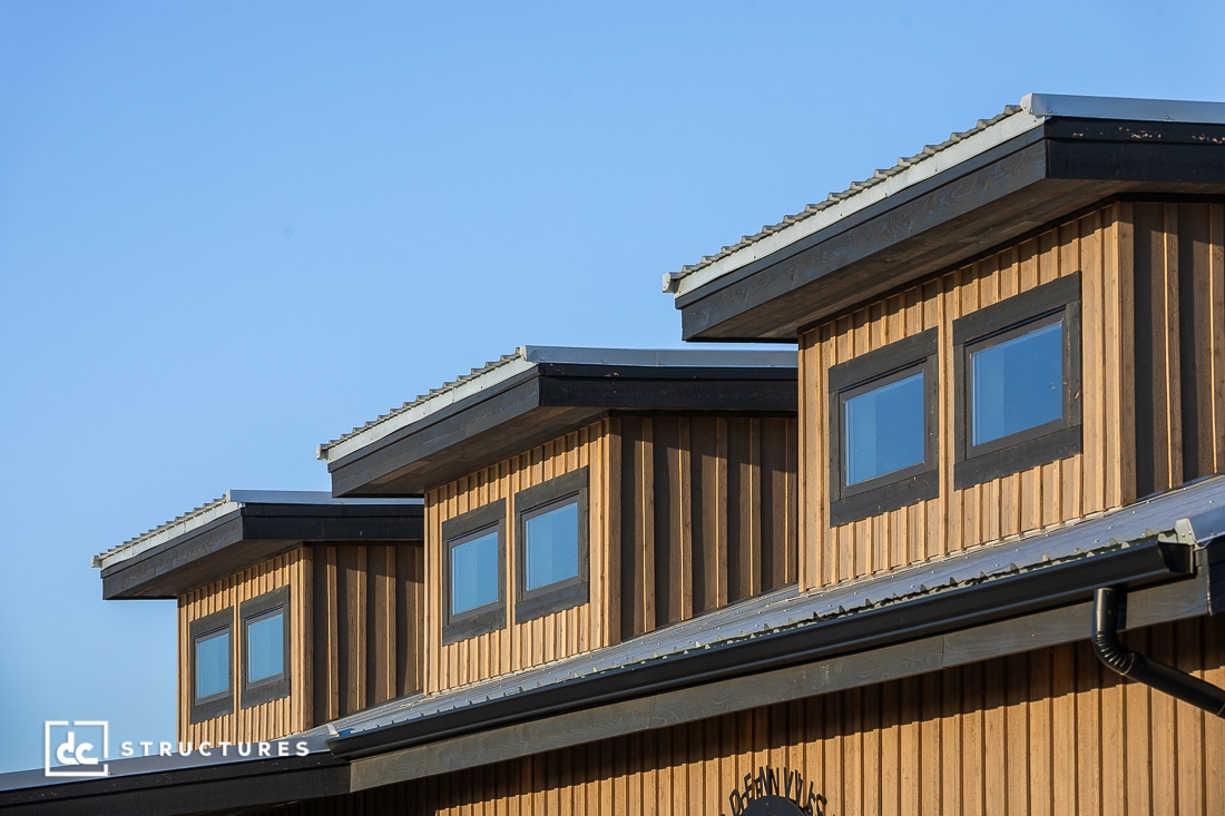 Wooden building with three rooftop dormers, each featuring two rectangular windows. Vertical wood paneling, black trim, clear blue sky.