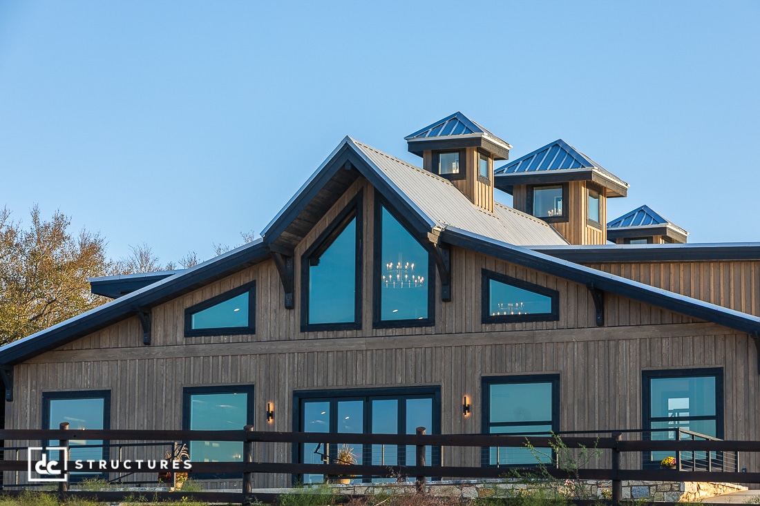 A modern barn-style building with large triangular windows, wood siding, a metal roof, and cupolas against a clear blue sky. A black fence runs in front of the building.