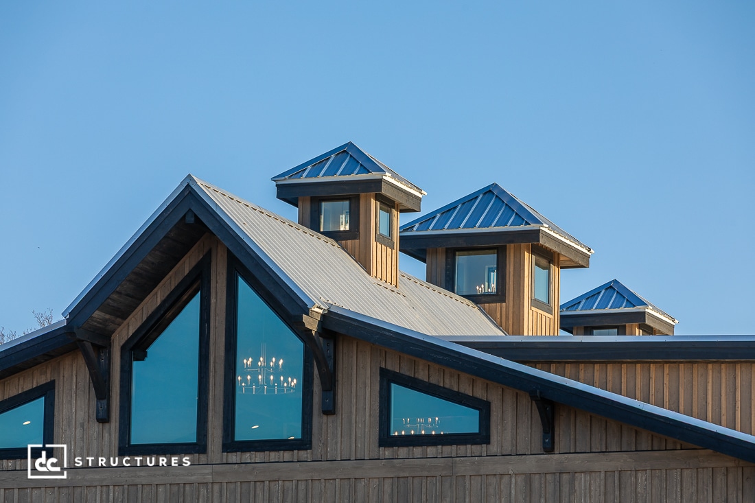 The image shows the upper part of a wooden barn-style building with blue metal roofs, three cupolas, large windows, and a chandelier inside. The sky is clear and blue.