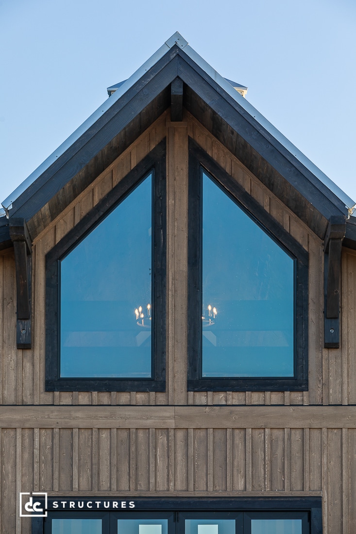 A close-up of the upper facade of a modern wooden barn with a steep gable roof and large triangular windows reflecting the sky; a chandelier is visible inside.