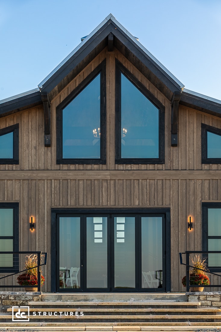 A modern wooden barn-style house with large triangular windows, black-framed glass doors, and symmetrical lighting fixtures. Potted plants border the steps leading to the entrance.