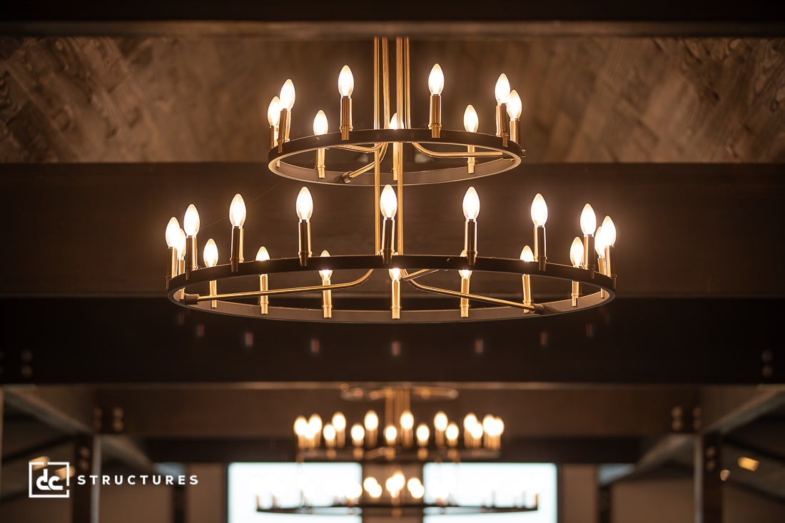 A close-up of a modern, double-tiered chandelier with lit candle-like bulbs, hanging from a ceiling with wooden beams.