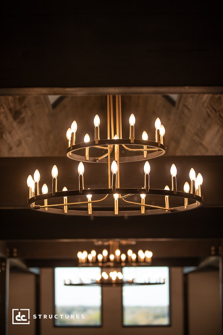 A modern two-tiered gold chandelier with lit candle-style bulbs hangs from a vaulted wooden ceiling, reflecting in a large window.
