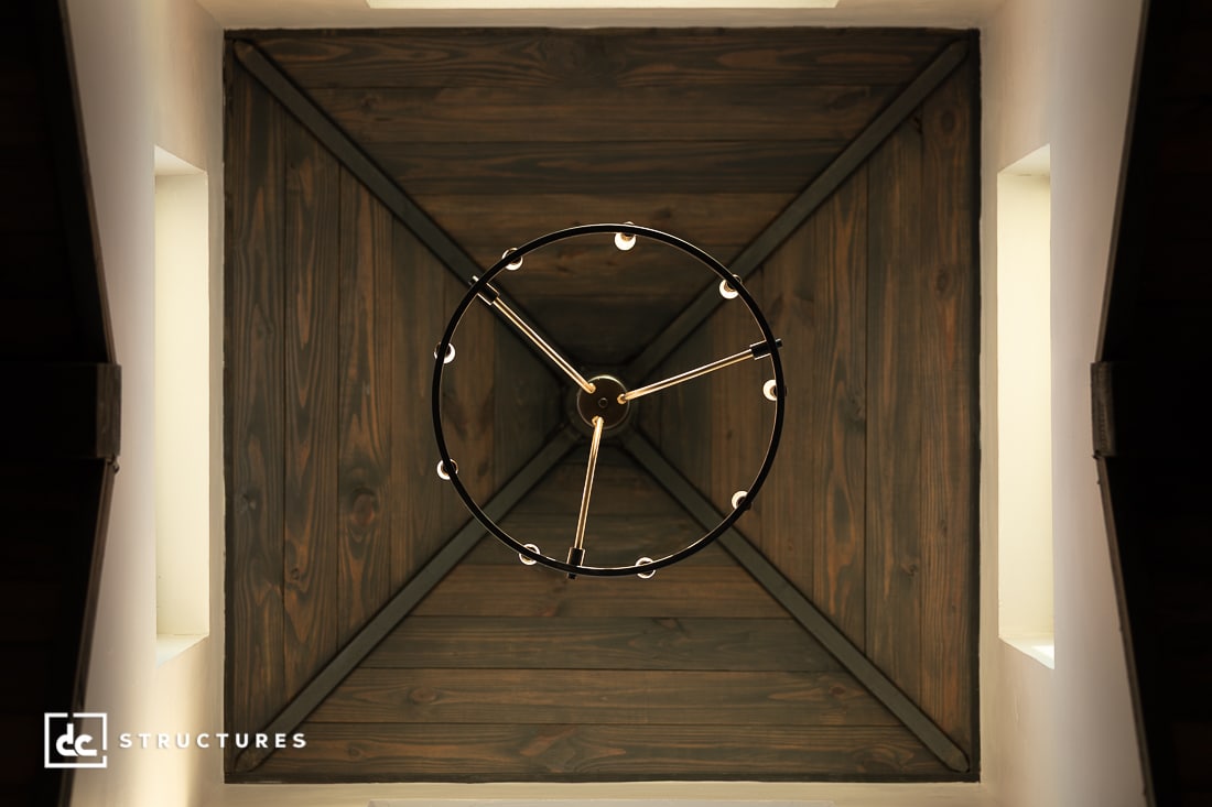 A circular chandelier with exposed bulbs hangs from a wooden, pyramid-shaped ceiling, viewed from below to highlight its symmetry.