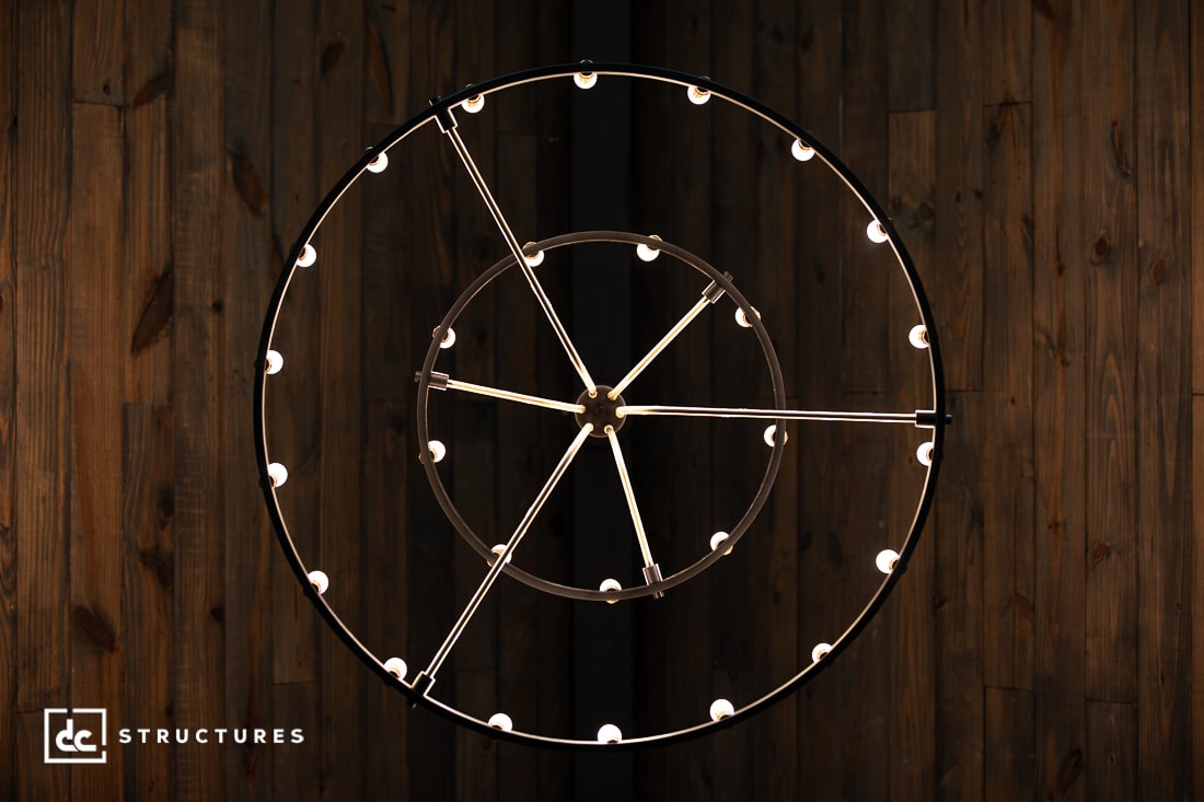A modern circular chandelier with multiple light bulbs is suspended against a dark, wooden plank wall.