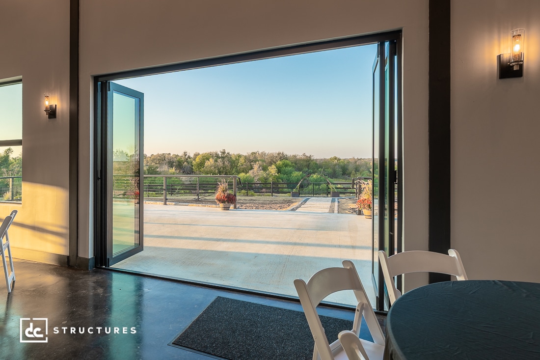 View from inside a modern room with glass folding doors open to a spacious patio, scenic landscape, white chairs and table visible.