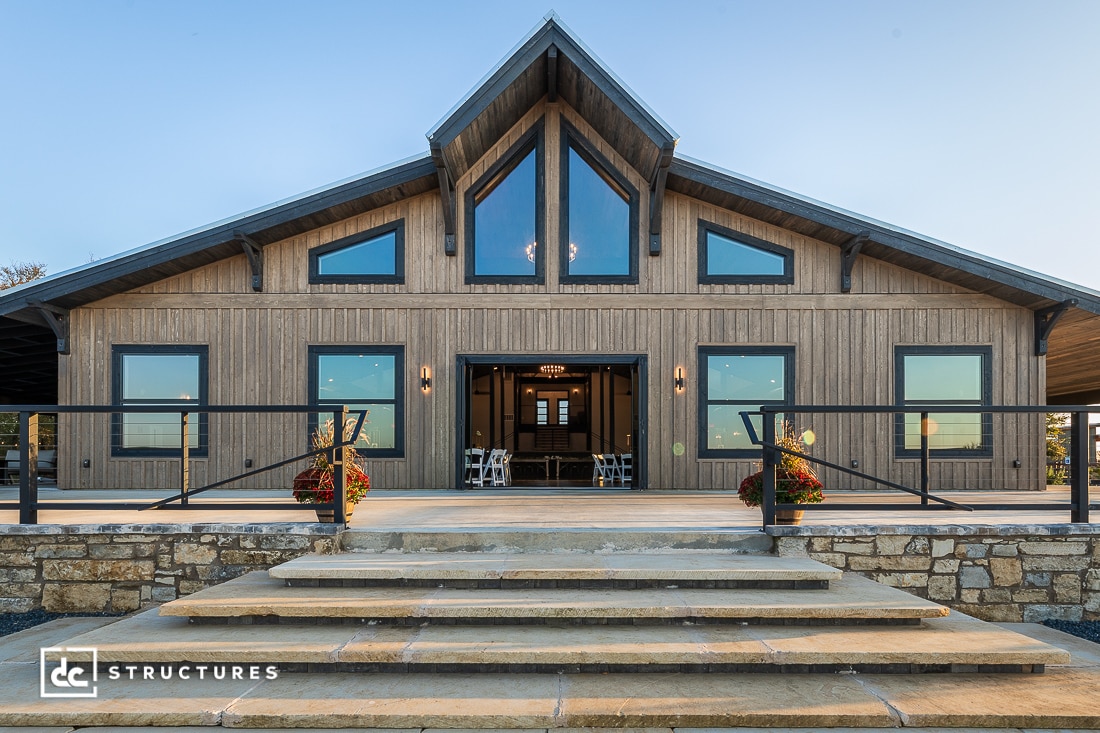 Modern barn-style building with wood siding, large glass windows, and a peaked roof. Stone steps lead up to the entrance, flanked by potted plants.