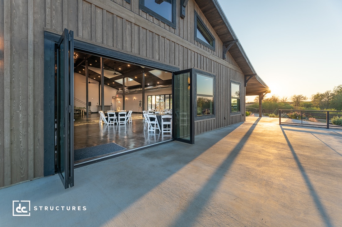 A modern barn-style building with open glass doors shows an event space with white chairs and tables as sunset casts warm light outside.