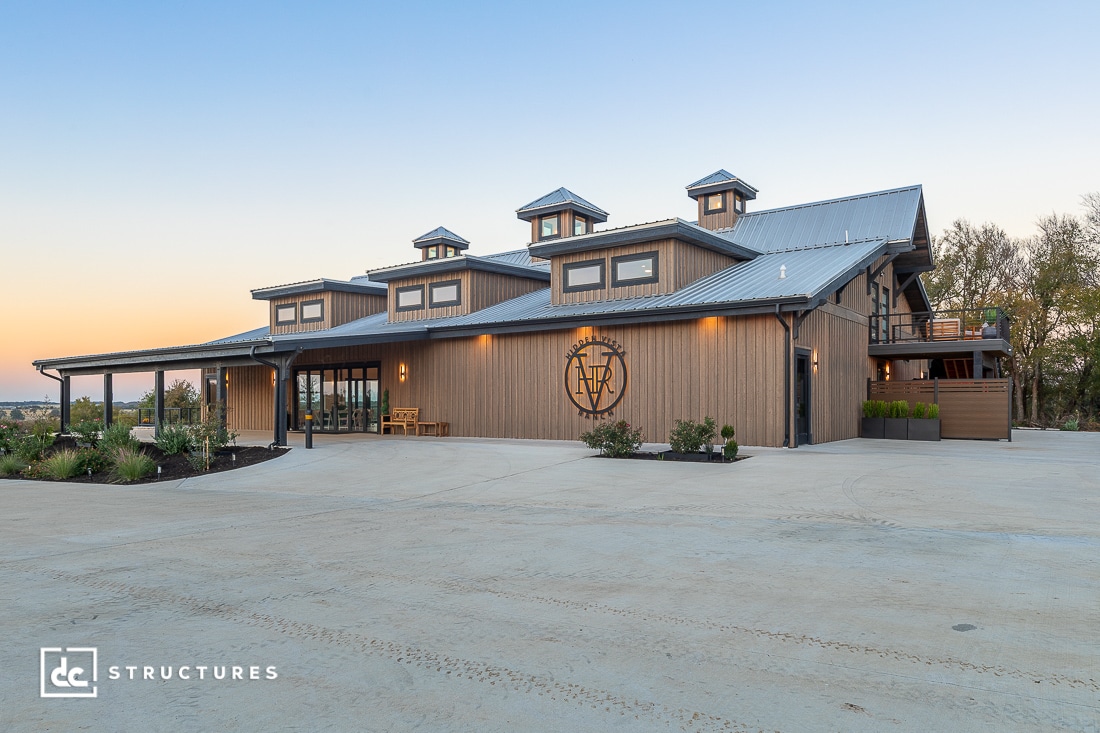 A large modern barn with a metal roof, wooden siding, and cupolas at sunset. Features porch, big windows, and landscaping.