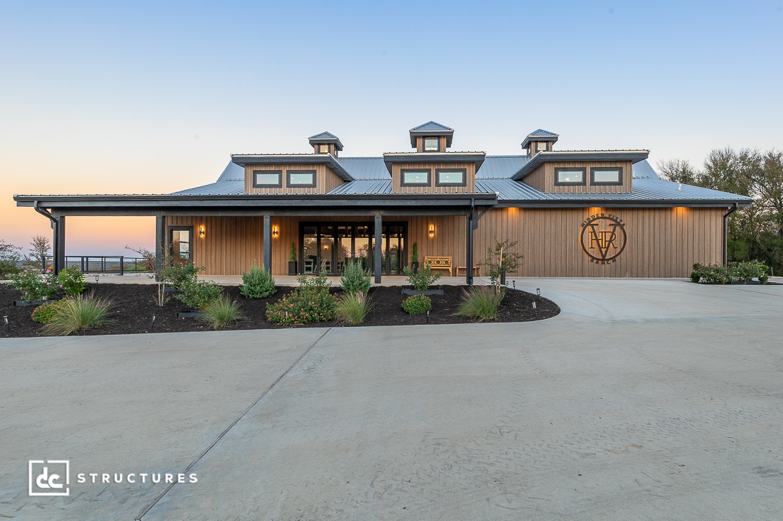A large modern barn-style building with wood siding, metal roof, and three cupolas, surrounded by landscaped plants at sunset.