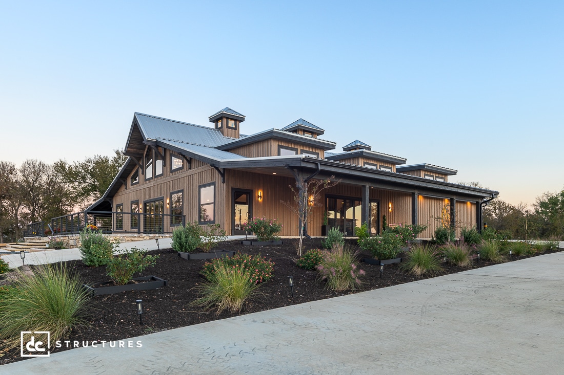 A modern barn-style building with wood siding, large windows, and cupolas, surrounded by plants and a concrete driveway at sunset.