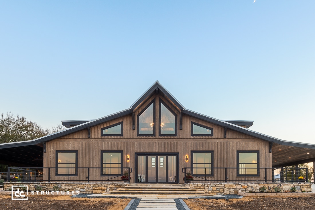 A modern barn-style house with large windows, wood siding, and a stone walkway to double front doors under a clear sky.