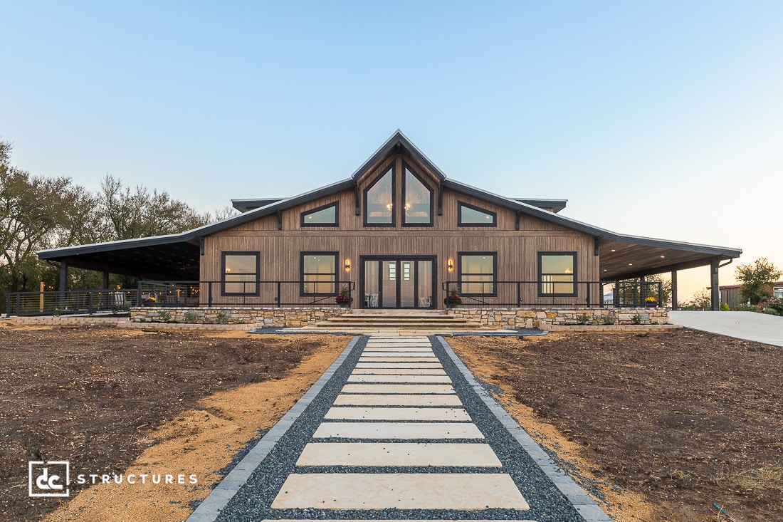 A modern barn-style house with large windows, a stone foundation, and a covered porch. A stone walkway leads to the entrance.