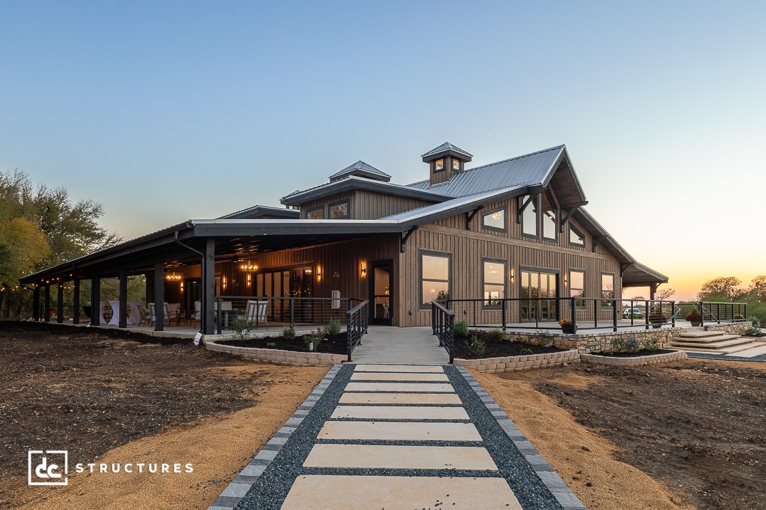 Modern barn-style building with large windows, a wraparound porch, and metal roof, surrounded by garden beds at sunset.