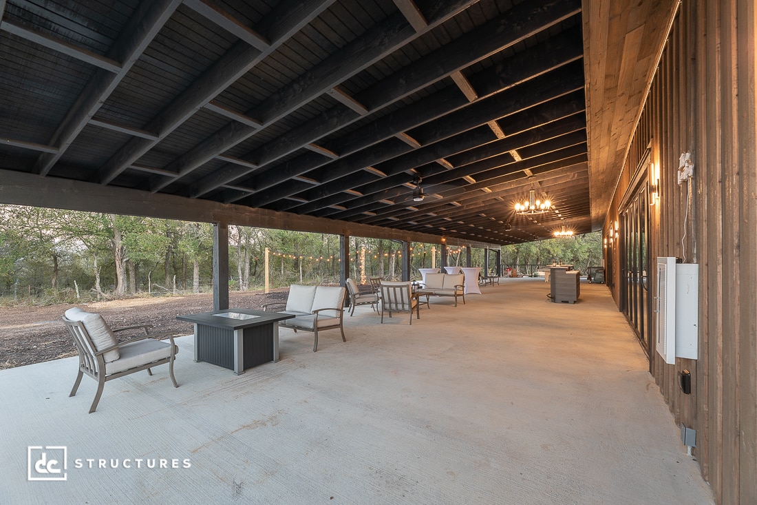 Spacious covered patio with outdoor seating, chandeliers, and a wooden ceiling; overlooks a natural landscape with trees.