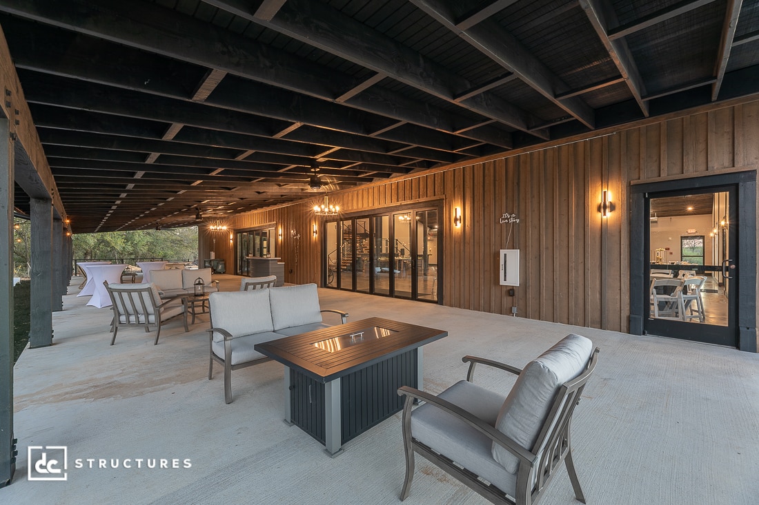 Covered outdoor patio with wood paneling, glass doors, and multiple seating areas, including cushioned chairs around a fire table.