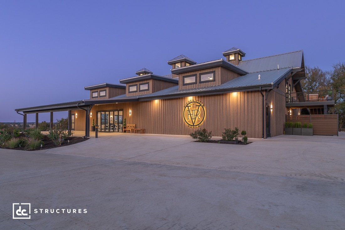 A large, modern barn-style building with wood siding, large windows, multiple cupolas, and warm outdoor lighting at dusk. Landscaped surroundings.