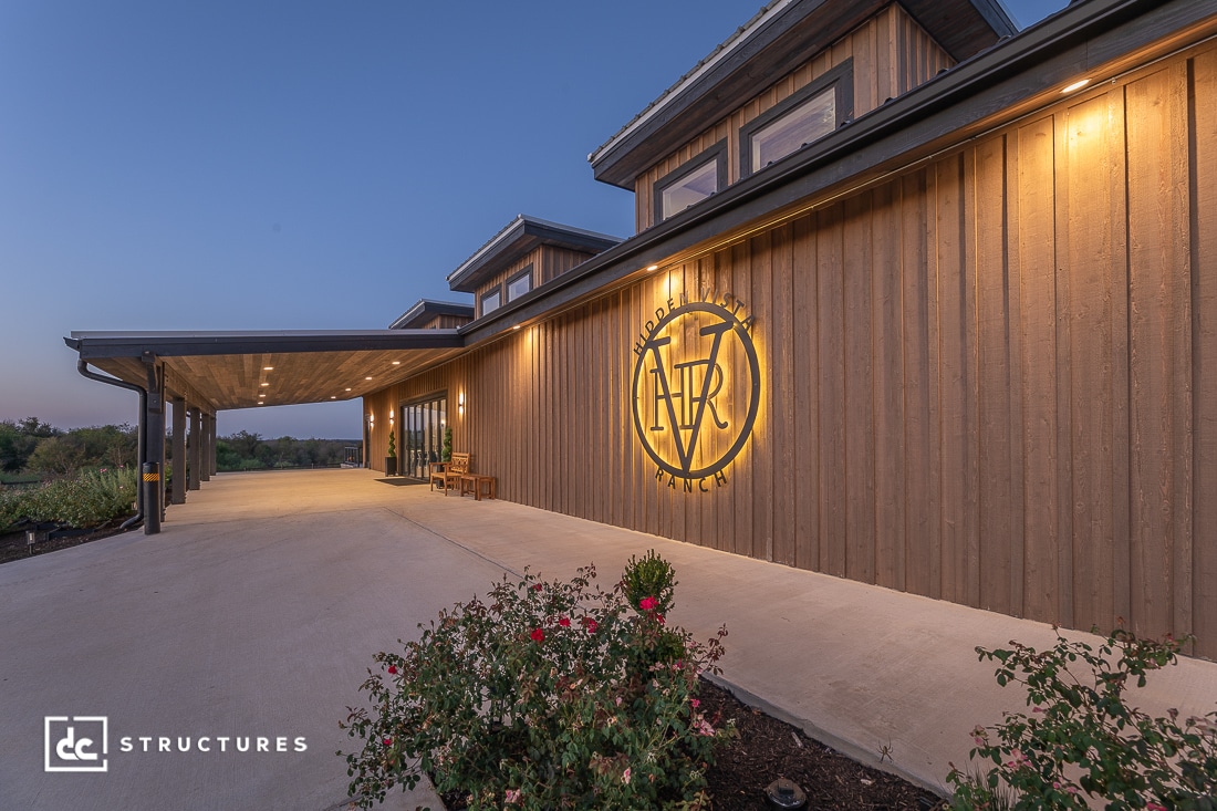 A modern wooden building with a large covered entryway and illuminated exterior lights at dusk. Bushes and flowers line the sidewalk.
