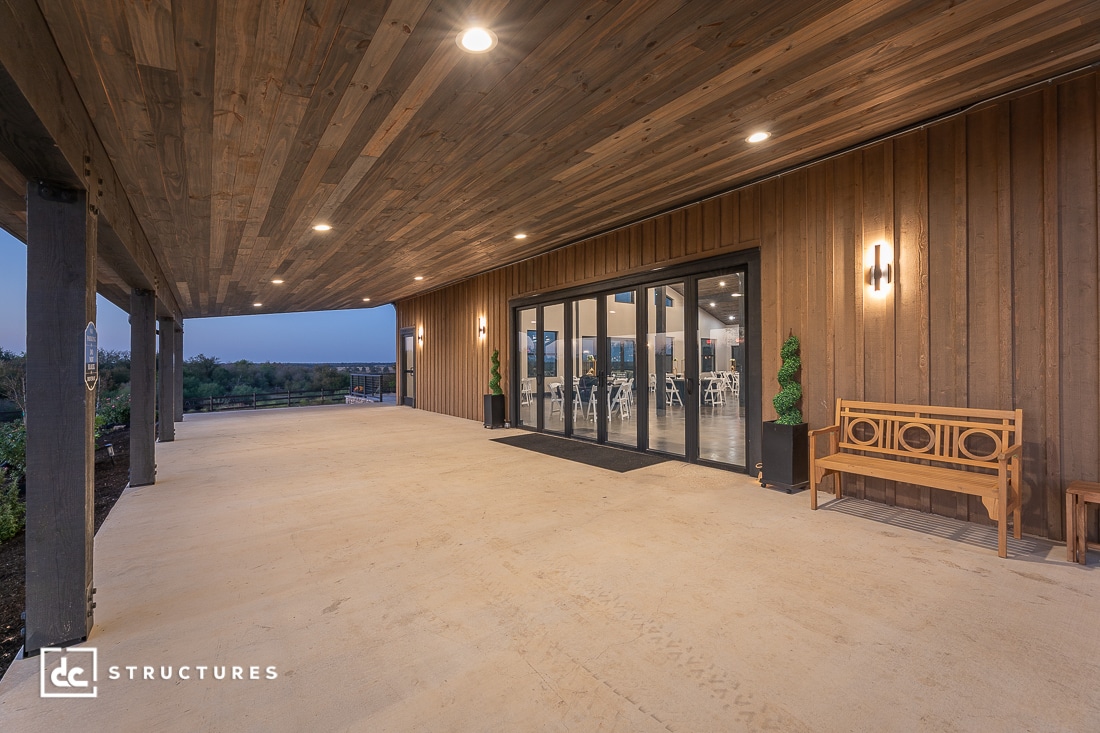 A covered outdoor patio with wooden ceiling and walls, modern lighting, a wooden bench, and large glass doors overlooking landscaping.