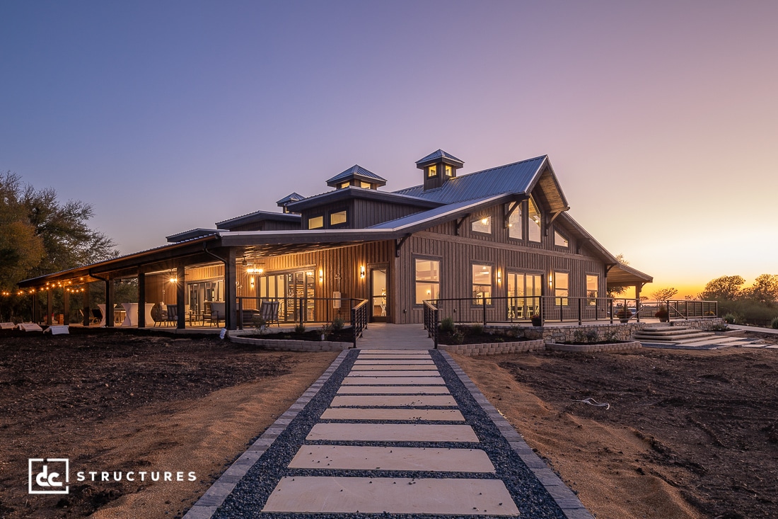 A modern barn-style building with large windows and a covered porch at sunset. Stone walkway leads to entrance, minimal vegetation.