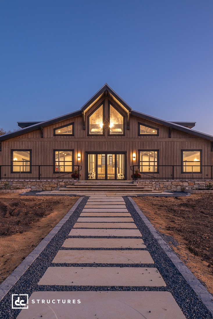 Modern barn-style house at dusk with warm lights, large windows, wooden siding, stone path entrance, clear sky, unfinished landscaping.