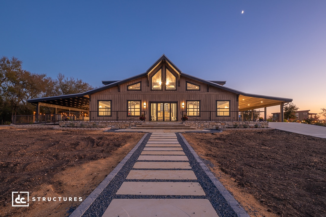 A modern barn-style house with large windows, warm lights at dusk, stone pathway, bare soil, and a crescent moon in the clear sky.