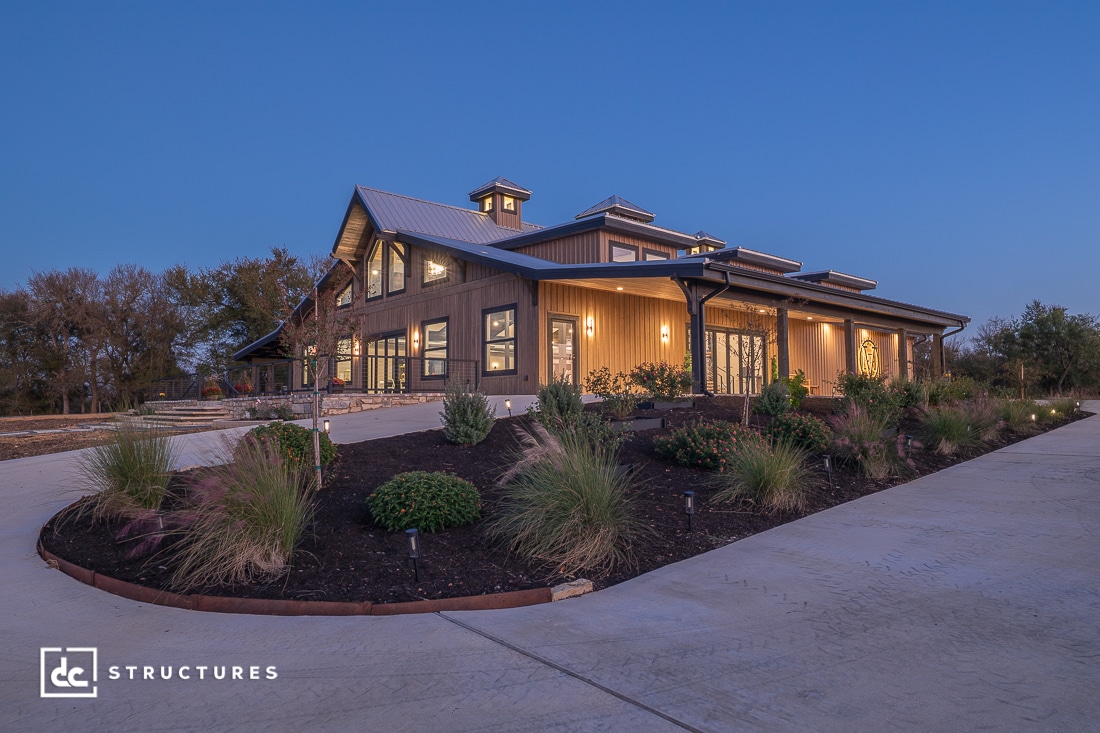 A modern barn-style house with large windows, outdoor lighting, and gardens sits by a wide concrete driveway at sunset.