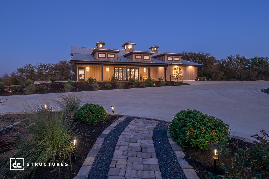 A large modern barn-style building with three cupolas is warmly lit at dusk, with a walkway and landscaping leading to the entrance.