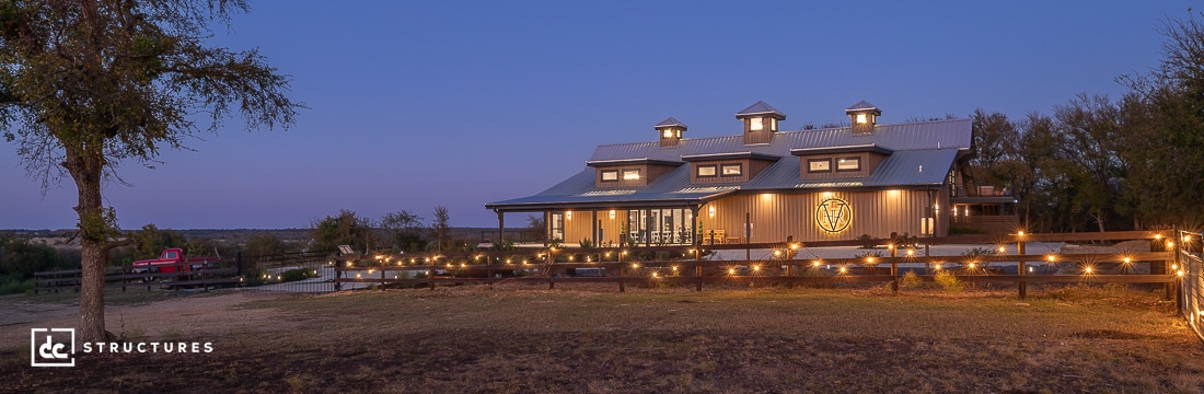 A large modern barn with cupolas is lit by string lights at dusk, surrounded by trees, a wooden fence, pond, and a red truck.