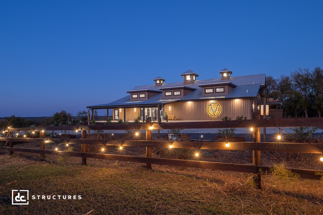 A modern barn-style building with large windows and cupolas is illuminated at dusk. String lights line a wooden fence in front.