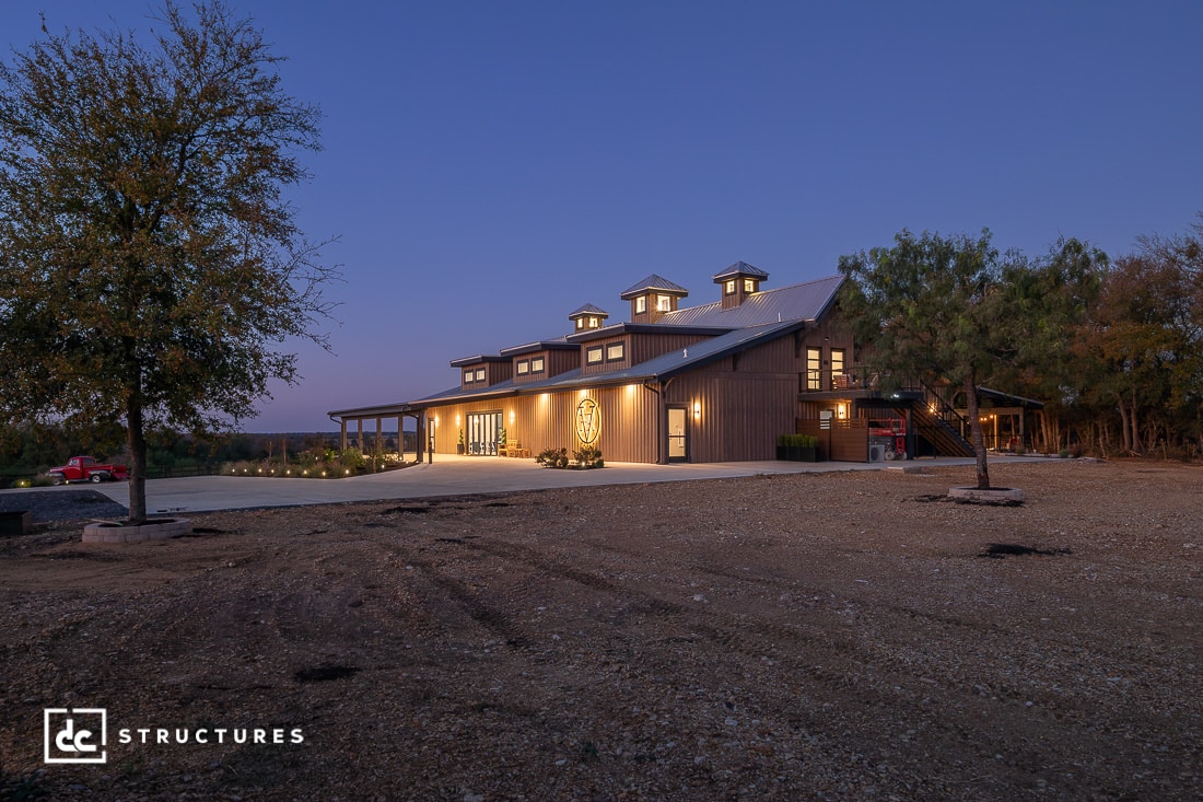 A large, modern barn-style building with warm exterior lights and multiple cupolas stands on a gravel lot at dusk, surrounded by trees. The sky is clear and deep blue.