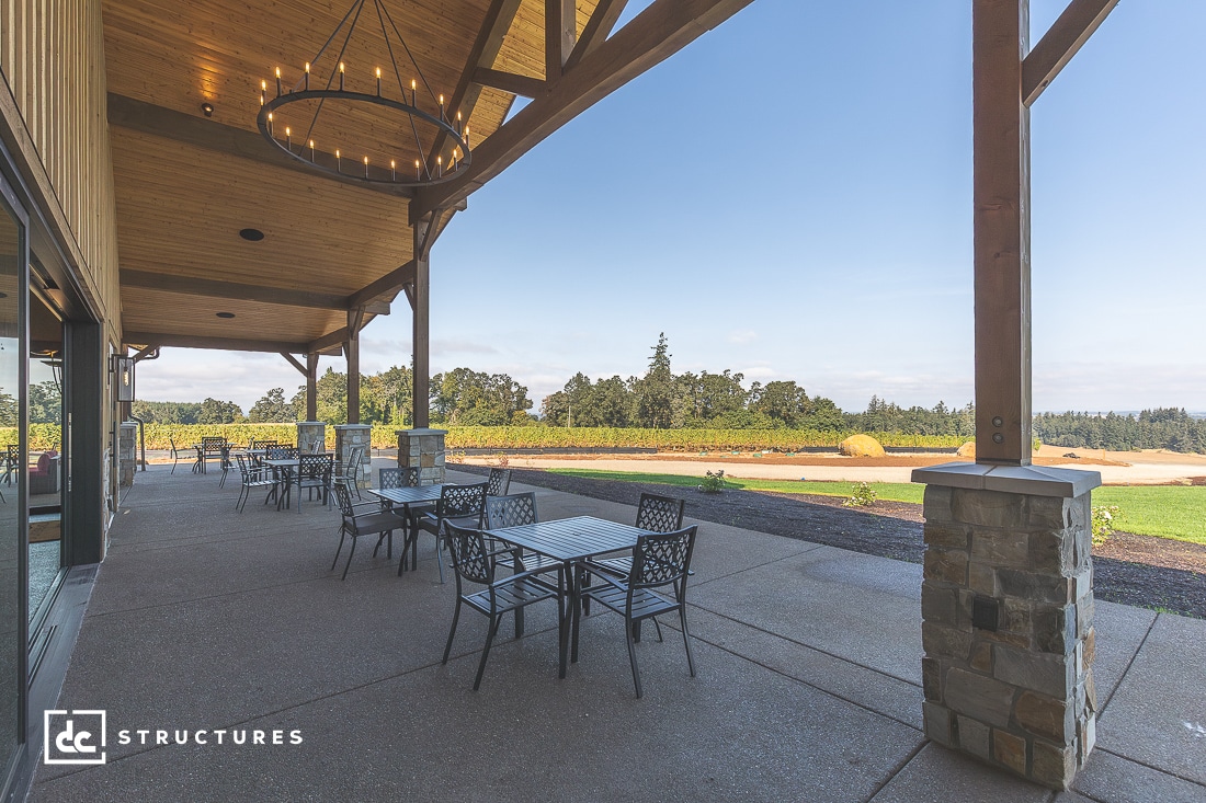 Covered outdoor patio with metal tables and chairs, stone pillars, wooden beams, overlooking a landscaped lawn and open fields.