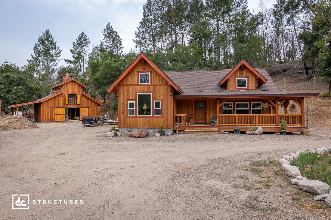 A rustic wooden house with a covered porch and large windows in a rural wooded area, with a barn and dirt driveways nearby.