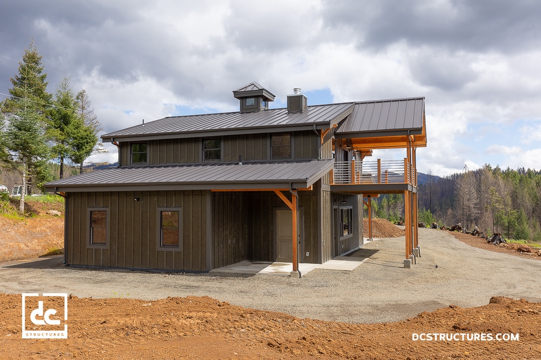 A modern two-story house with dark wood siding, metal roof, and a covered balcony, surrounded by trees and a gravel driveway.