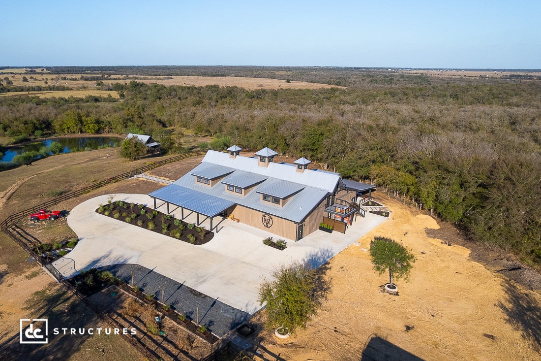 Aerial view of a large modern barn with a metal roof, surrounded by open fields, trees, a pond, a red vehicle, and a paved driveway.