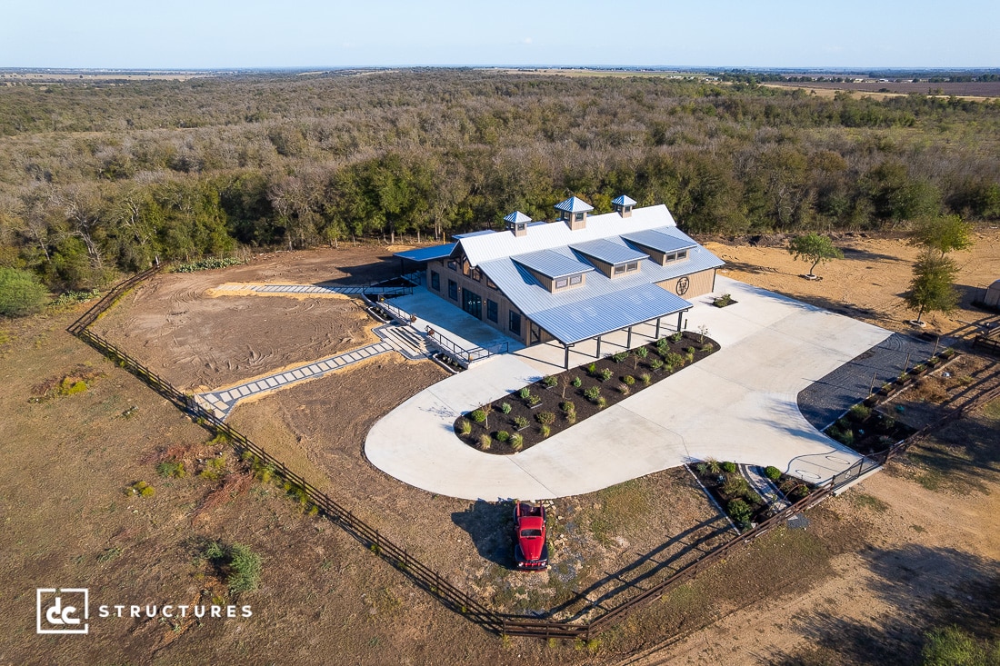 Aerial view of a modern barn with a metal roof, curved driveway, landscaped areas, fencing, open fields, and a red vehicle nearby.