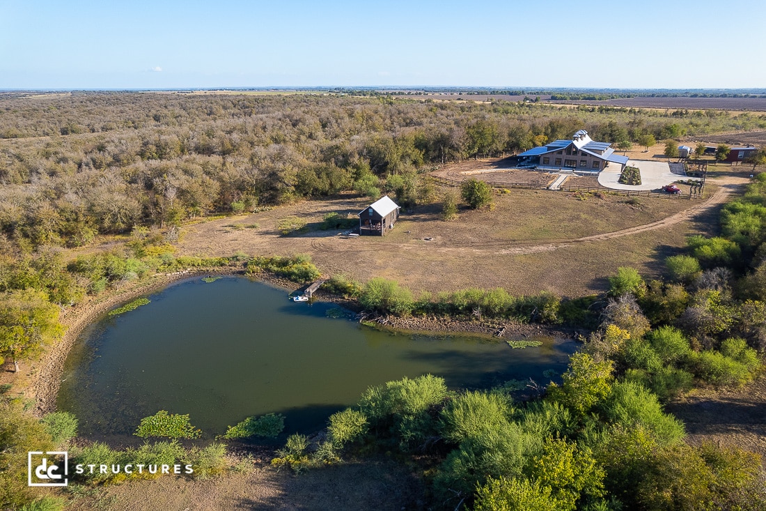 Aerial view of a rural property with a pond, small barn, and large house surrounded by trees and open fields under a clear sky.