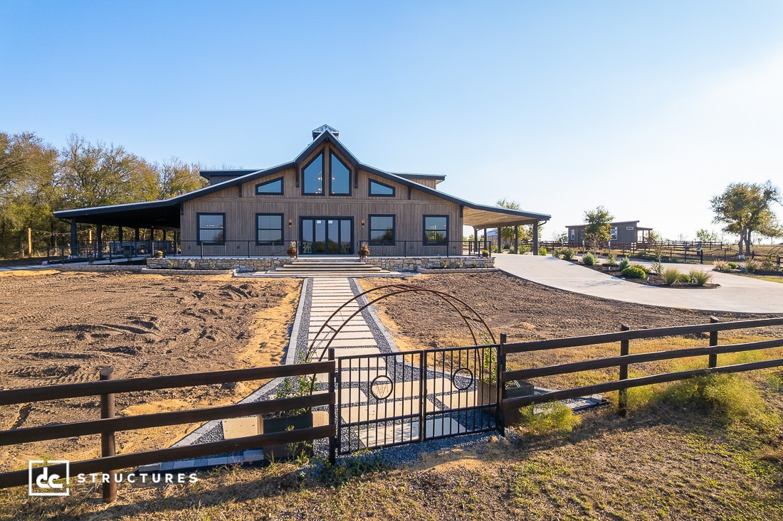 A modern barn-style house with large windows sits behind a black gate and wooden fence, with a stone walkway to the entrance.